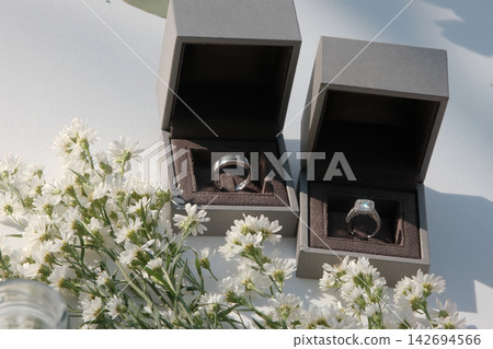 The wedding rings are in boxes on a corner of a white table, decorated with dried flowers The wedding rings are in boxes on a corner of a white table, decorated with dried flowers 142694566