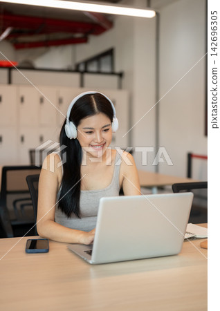 Young Asian woman wearing headphones smiling while typing on laptop at table in a co working cafe. 142696305