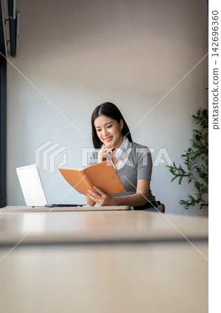 Young Asian woman reading a book while sitting beside a laptop at table in a co working cafe. 142696360