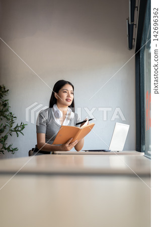 Young Asian woman looking away from book while sitting beside a laptop at table in a co working cafe 142696362