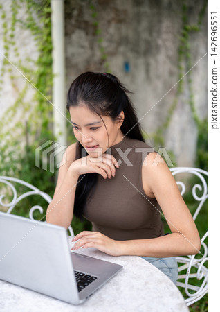 Focused Asian Woman Typing On Laptop At Outdoor Garden Cafe 142696551