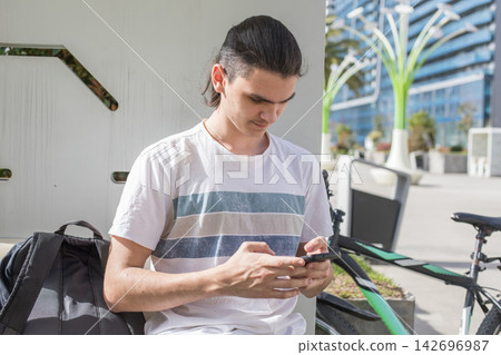A young man is seated on a bench looking at his phone with focus. A young man is seated on a bench looking at his phone with focus. 142696987
