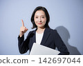 A smiling woman in her 50s in a suit against a light blue background, holding a laptop, in a business image, pointing to a specific point. 142697044