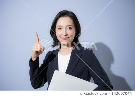 A smiling woman in her 50s in a suit against a light blue background, holding a laptop, in a business image, pointing to a specific point. A smiling woman in her 50s in a suit against a light blue background, holding a laptop, in a business image, pointing to a specific point. 142697044