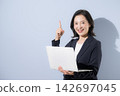 A smiling woman in her 50s in a suit against a light blue background, holding a laptop, in a business image, pointing to a specific point. 142697045