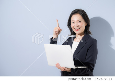 A smiling woman in her 50s in a suit against a light blue background, holding a laptop, in a business image, pointing to a specific point. A smiling woman in her 50s in a suit against a light blue background, holding a laptop, in a business image, pointing to a specific point. 142697045