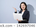 A smiling woman in her 50s in a suit against a light blue background, holding a laptop; a business image, suggesting a recommendation or introduction pose. 142697046