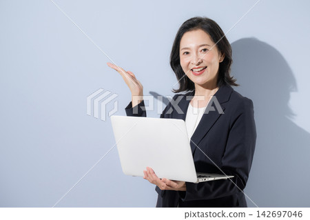 A smiling woman in her 50s in a suit against a light blue background, holding a laptop; a business image, suggesting a recommendation or introduction pose. A smiling woman in her 50s in a suit against a light blue background, holding a laptop; a business image, suggesting a recommendation or introduction pose. 142697046