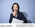 A smiling woman in her 50s in a suit with a light blue background, holding a laptop – a business image that is surprising and astonishing. 142697293