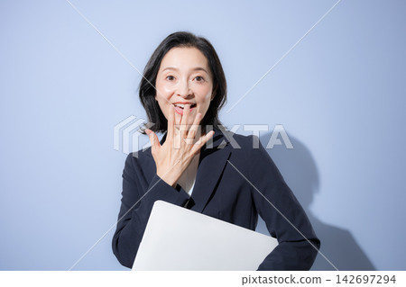 一位 50 多歲、微笑的女士身穿淺藍色背景的西裝,手持筆記型電腦——這是一張令人驚訝和震撼的商務照片。 一位 50 多歲、微笑的女士身穿淺藍色背景的西裝,手持筆記型電腦——這是一張令人驚訝和震撼的商務照片。 142697294