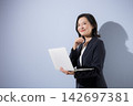 A smiling woman in her 50s, wearing a suit against a light blue background and holding a laptop, is shown in a smiling, thoughtful, or troubled pose, perhaps representing a business career. 142697381