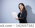 A smiling woman in her 50s, wearing a suit against a light blue background and holding a laptop, strikes a pose that suggests she is seriously contemplating business. 142697382