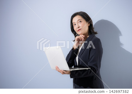 A smiling woman in her 50s, wearing a suit against a light blue background and holding a laptop, strikes a pose that suggests she is seriously contemplating business. A smiling woman in her 50s, wearing a suit against a light blue background and holding a laptop, strikes a pose that suggests she is seriously contemplating business. 142697382