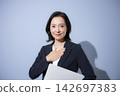 A smiling woman in her 50s, wearing a suit against a light blue background and holding a laptop, strikes a pose that conveys business confidence, reliability, and peace of mind, while looking directly at the camera. 142697383