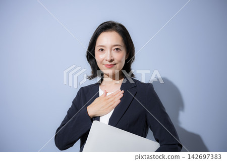 A smiling woman in her 50s, wearing a suit against a light blue background and holding a laptop, strikes a pose that conveys business confidence, reliability, and peace of mind, while looking directly at the camera. A smiling woman in her 50s, wearing a suit against a light blue background and holding a laptop, strikes a pose that conveys business confidence, reliability, and peace of mind, while looking directly at the camera. 142697383