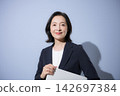 A smiling woman in her 50s in a suit against a light blue background, holding a laptop; a business image, representing working women, career changes, and career development. 142697384