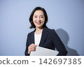 A smiling woman in her 50s in a suit against a light blue background, holding a laptop; a business image, representing working women, career changes, and career development. 142697385