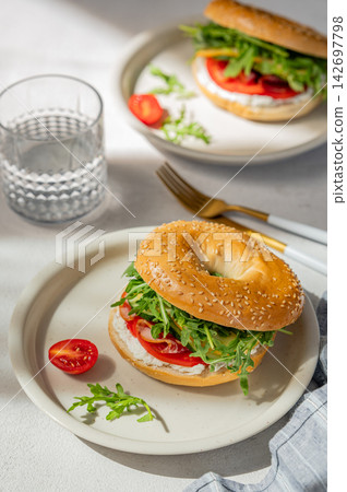 Two fresh bagels with tomato, prosciutto, cheese and arugula on a plates on a light background Two fresh bagels with tomato, prosciutto, cheese and arugula on a plates on a light background 142697798