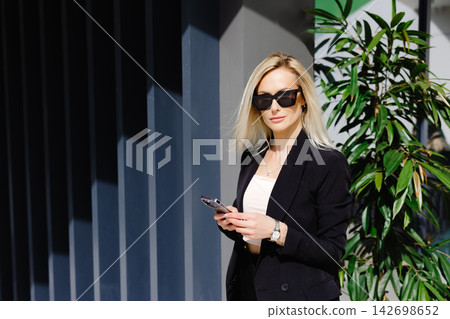 Young businesswomen in black suit using smartphone isolated on background gray wall. Pretty manager holding mobile gadget and texting message Young businesswomen in black suit using smartphone isolated on background gray wall. Pretty manager holding mobile gadget and texting message 142698652