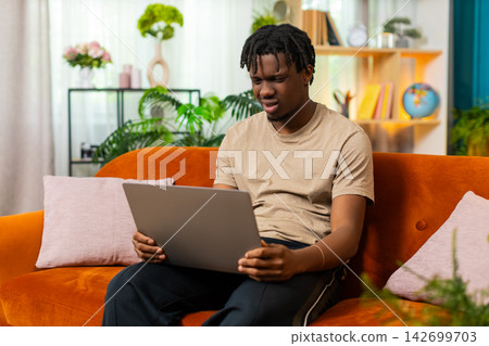African American young man on home sofa upset by laptop error and sighing during online work task African American young man on home sofa upset by laptop error and sighing during online work task 142699703