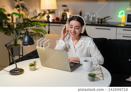 Young woman works on laptop wearing wireless headphones types slow writes plan for tomorrow evening Young woman works on laptop wearing wireless headphones types slow writes plan for tomorrow evening 142699728