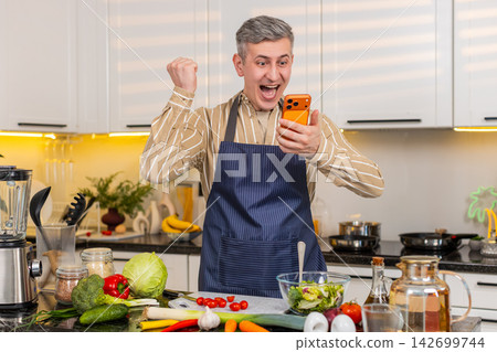 Middle-aged man celebrating victory with smartphone in kitchen after online success at home smiling Middle-aged man celebrating victory with smartphone in kitchen after online success at home smiling 142699744