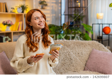 Young woman on home sofa checking phone and cash while planning monthly home budget with calm smile Young woman on home sofa checking phone and cash while planning monthly home budget with calm smile 142699807