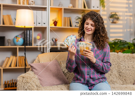 Young woman on home sofa counting cash and smiling after salary bonus win or debt repayment indoors 142699848
