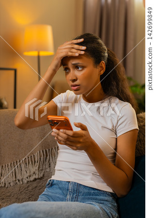 African American young woman on home sofa shows disappointment after reading bad news on smartphone African American young woman on home sofa shows disappointment after reading bad news on smartphone 142699849