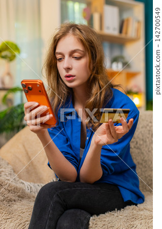 Young woman on home sofa paying online purchase with smartphone and bank card smiling at home calm Young woman on home sofa paying online purchase with smartphone and bank card smiling at home calm 142700549