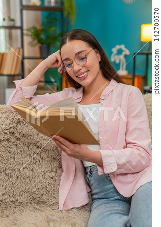 Young woman reading book on sofa turning pages slowly reflecting deeply on story absorbing meaning 142700570