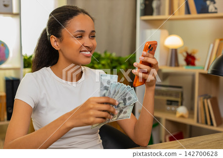 African American young woman on home table counting cash and checking personal budget on smartphone African American young woman on home table counting cash and checking personal budget on smartphone 142700628