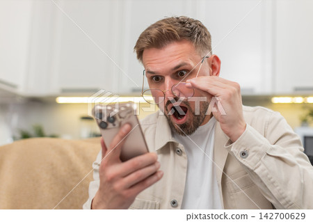 Young man with glasses reads small text on smartphone amazed by story fresh alert brings wide smile Young man with glasses reads small text on smartphone amazed by story fresh alert brings wide smile 142700629