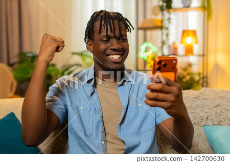 African American young man on home sofa celebrating cash message and online game win on smartphone 142700630