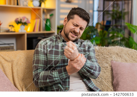 Young man on home sofa feeling wrist pain and doing wrist stretch exercise for pain relief indoors Young man on home sofa feeling wrist pain and doing wrist stretch exercise for pain relief indoors 142700633