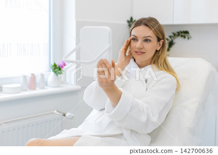Caucasian young woman smiles in mirror after facial therapy in beauty clinic showing visible result 142700636