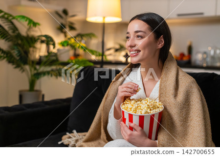 Young woman wrapped in plaid holds popcorn watches comedy on tv laughs comments comfy cozy evening Young woman wrapped in plaid holds popcorn watches comedy on tv laughs comments comfy cozy evening 142700655