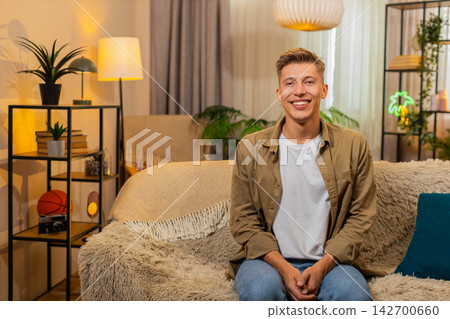 Young man on home sofa sitting straight and smiling at camera in calm portrait in cozy home indoors Young man on home sofa sitting straight and smiling at camera in calm portrait in cozy home indoors 142700660