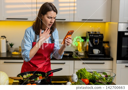 Young woman shocked by smartphone message in home kitchen feeling defeat and disappointment indoors Young woman shocked by smartphone message in home kitchen feeling defeat and disappointment indoors 142700663