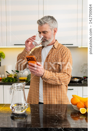 Middle-aged man checks smartphone while drinking water in kitchen for healthy diet hydration routine Middle-aged man checks smartphone while drinking water in kitchen for healthy diet hydration routine 142700664