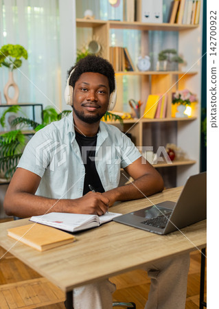 African American young man on home table studying with headphones laptop and notes for exams indoors African American young man on home table studying with headphones laptop and notes for exams indoors 142700922