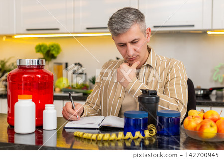 Middle-aged man writing fitness and nutrition plan in notebook for wellness routine in home kitchen 142700945