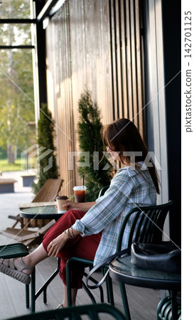 Relaxed Woman Sitting at an Outdoor Cafe Terrace with Takeaway Coffee on a Warm Summer Day Relaxed Woman Sitting at an Outdoor Cafe Terrace with Takeaway Coffee on a Warm Summer Day 142701125