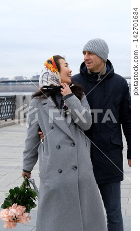 Two adults are kissing on a quiet shore. The woman is holding pale pink flowers and wearing a patterned headscarf. Soft, overcast light, a blurred shoreline, an open sky, and a serene city skyline. Two adults are kissing on a quiet shore. The woman is holding pale pink flowers and wearing a patterned headscarf. Soft, overcast light, a blurred shoreline, an open sky, and a serene city skyline. 142701684