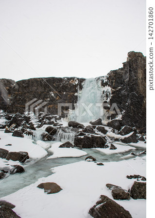 Vertical landscape of a partially frozen waterfall cascading over dark basalt cliffs with snow in Iceland 142701860