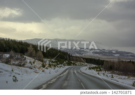 Empty asphalt road winding through a winter forest landscape with snow-capped mountains under a moody sky in Iceland. Empty asphalt road winding through a winter forest landscape with snow-capped mountains under a moody sky in Iceland. 142701871