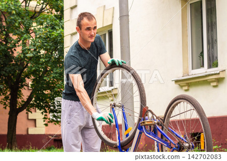 Man fixing bicycle wheel at doorstep. Own vehicle maintenance without service center. Responsible ownership visible. Summer commute switched to pedal power 142702083