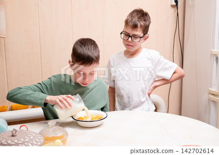 A young boy pouring fresh milk from a glass jar into a bowl of cereal at the kitchen table, while his brother in glasses stands next to him A young boy pouring fresh milk from a glass jar into a bowl of cereal at the kitchen table, while his brother in glasses stands next to him 142702614