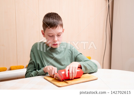 A young caucasian boy in a green long-sleeved shirt carefully cutting a big red sausage with a knife on a wooden chopping board at home. A young caucasian boy in a green long-sleeved shirt carefully cutting a big red sausage with a knife on a wooden chopping board at home. 142702615