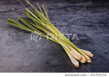 Fresh lemongrass stalks with green leaves on dark textured surface, natural lighting, shallow depth of field, copy space Fresh lemongrass stalks with green leaves on dark textured surface, natural lighting, shallow depth of field, copy space 142704158